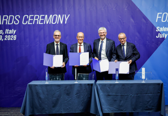 Four individuals stand at a table holding open blue folders during an awards ceremony in Salerno, Italy, on July 19, 2026.