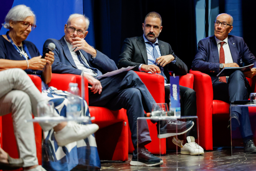 Four individuals are seated on a stage in red chairs during a panel discussion, with one person holding a microphone and another holding papers.